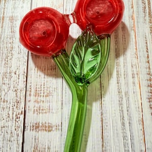 May include: A glass pipe shaped like two red cherries on a single green stem. The two red circular bowls are connected at the top, and a green glass leaf is attached to the stem. The stem is long and cylindrical, serving as the mouthpiece for the pipe.