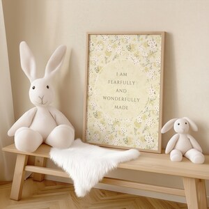 May include: A white wooden bench with a white faux fur throw on top. A white stuffed bunny sits on the bench. A framed print with a floral design and the text "I am fearfully and wonderfully made" hangs on the wall behind the bench.