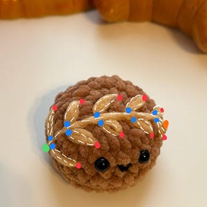 May include: A brown crocheted plush toy with a smiling face and black eyes. The toy has a gold crocheted wreath with white stitching around its head.