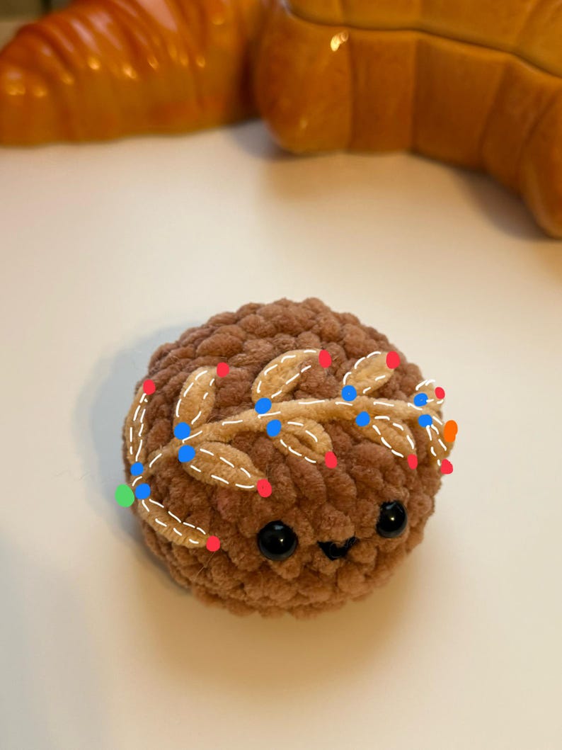 May include: A brown crocheted plush toy with a smiling face and black eyes. The toy has a gold crocheted wreath with white stitching around its head.