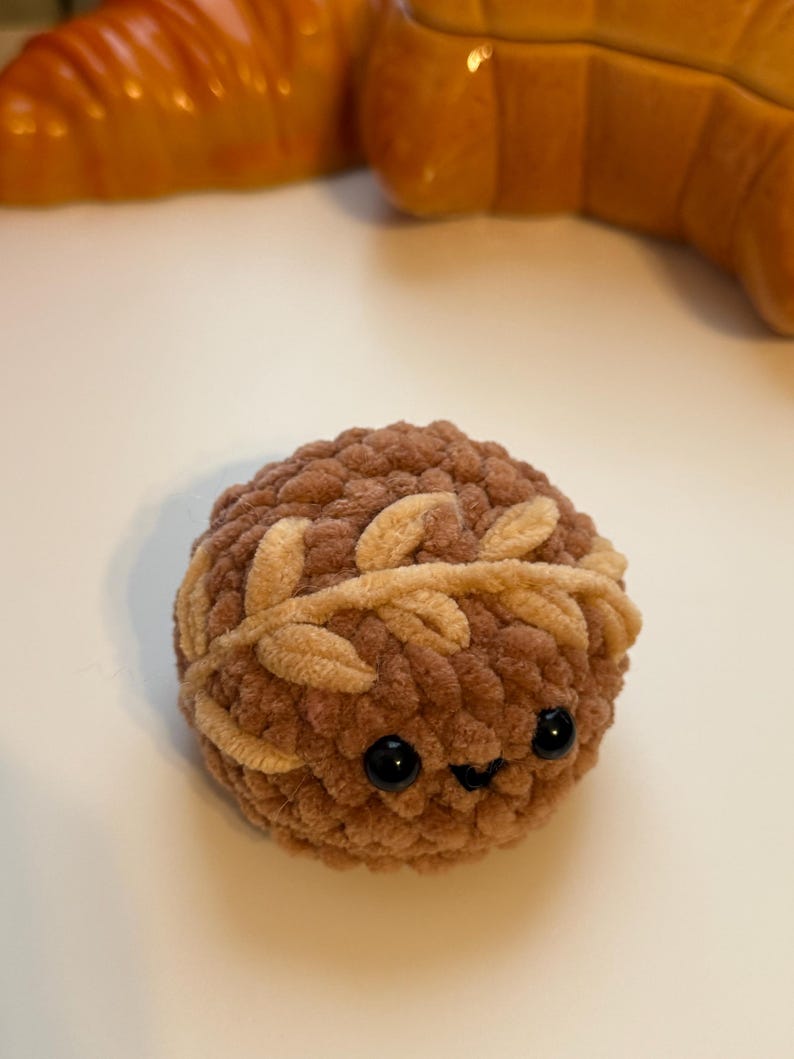 May include: A brown crocheted plush toy with a smiling face and a brown and beige crocheted wreath around its head.