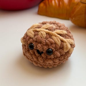 May include: A brown crocheted plush toy shaped like a bun with a smiling face and black eyes. The bun has a brown and beige crocheted design.