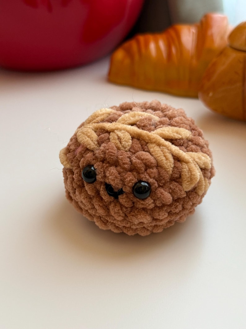 May include: A brown crocheted plush toy shaped like a bun with a smiling face and black eyes. The bun has a brown and beige crocheted design.