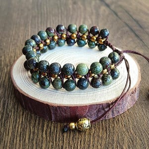 Op de afbeelding: Een armband met twee rijen groene en bruine jaspis kralen en goudkleurige metalen kralen. De armband is gebonden met bruin koord en heeft een goudkleurige metalen kraal aan het einde.