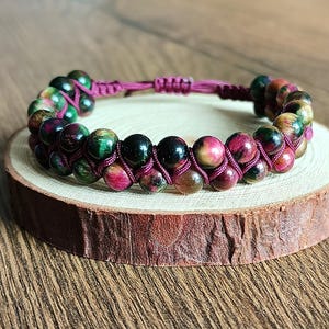 Op de afbeelding: Een armband met dubbele strengen met veelkleurige kralen in groen, rood en bruin. De kralen zijn geregen op een donkerroze koord, waardoor een gevlochten effect ontstaat. De armband wordt gepresenteerd op een klein houten schijfje.