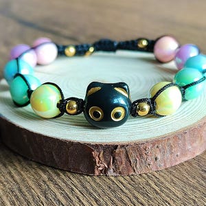 Op de afbeelding: Een kralenarmband met een zwarte kattenbedel en iriserende kralen. De kralen zijn in tinten roze, blauw, groen en geel, geregen op zwart koord met goudkleurige kralen. De armband wordt gepresenteerd op een houten schijf.