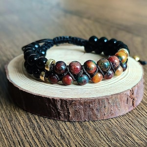 Può includere: Un braccialetto di perline nere e multicolori. Il braccialetto è realizzato con un cordoncino nero e presenta dettagli metallici color oro. Le perline sono nelle tonalità del rosso, verde, arancione e nero. Il braccialetto è esposto su una piccola fetta di legno.