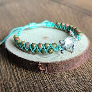 May include: A turquoise and green beaded bracelet with a silver turtle charm. The bracelet is tied with a turquoise cord and is displayed on a wooden surface.