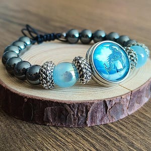 Può includere: Un braccialetto di perline con perline di ematite grigio scuro, perline iridescenti azzurre e una grande perlina focale rotonda con un disegno blu e bianco. Il braccialetto è esposto su una piccola fetta di legno.