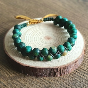 Op de afbeelding: Een groene kralenarmband met een macramé sluiting. De armband is gemaakt van ronde, donkergroene kralen en wordt gepresenteerd op een kleine houten schijf. De macramé sluiting is bruin en zwart.