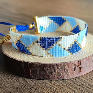 May include: A beaded bracelet featuring a diamond pattern in shades of blue and white, with gold-coloured metal details and a blue cord fastening. The bracelet is presented on a small, round wooden slice.
