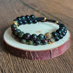 Op de afbeelding: Een armband met dubbele rij zwarte en groene kralen, geaccentueerd met gouden kralen. De armband is gemaakt van natuurlijke stenen en is vastgemaakt met een zwart koord.