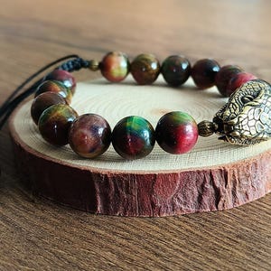 May include: A beaded bracelet with large, round beads in red, green, and brown. It has a brass dragon head clasp and a black adjustable cord. The bracelet is displayed on a small wooden slice. The beads are approximately 1 cm in diameter.