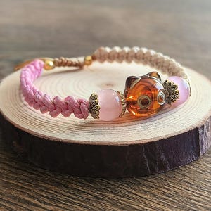 May include: A handcrafted bracelet with a pink braided cord and a tan macrame section. It features a clear orange cat-shaped bead, pink beads, and gold-coloured metal accents. The bracelet is displayed on a wooden slice.