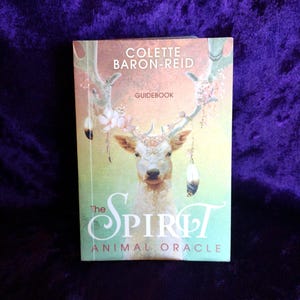 May include: A book titled "The Spirit Animal Oracle" by Colette Baron-Reid. The cover shows a deer with floral antlers and hanging feathers, set against a gradient background of peach, green, and blue.