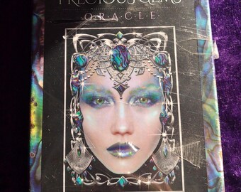 May include: A boxed oracle deck titled "Precious Gems Oracle" featuring a woman's face with elaborate gemstone makeup in shades of blue, green, and silver. The artist's name, Maxine Gadd, is printed at the bottom.