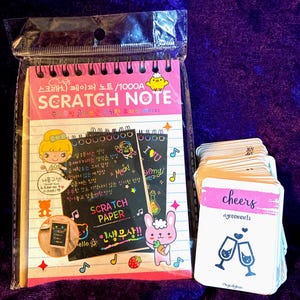 Pode incluir: Um bloco de notas de raspar com espiral numa capa de plástico transparente, com uma capa cor-de-rosa com ilustrações de desenhos animados e texto coreano, ao lado de um baralho de cartas brancas com a palavra cheers e dois copos.