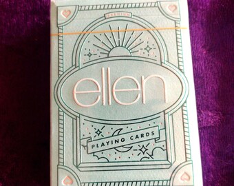 May include: A deck of "ellen" playing cards in a light blue box with an art deco design. The box features the word "ellen" in white, with the words "PLAYING CARDS" below. The deck is secured with a gold rubber band.