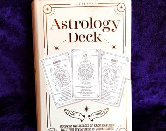 May include: A cream-colored Astrology Deck box with gold accents and the title "Astrology Deck" in elegant lettering. Three zodiac cards are displayed, featuring illustrations of Scorpio, Leo, and Pisces. The box is on a purple velvet surface.