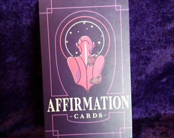 May include: A deck of affirmation cards in a rectangular box. The box is dark purple with the words "AFFIRMATION - CARDS-" in white. The design features a pink figure within a circular frame, with stars and lines.