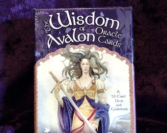 May include: A deck of "The Wisdom of Avalon Oracle Cards" by Colette Baron-Reid. The box features an illustration of a woman holding a sword, with text that reads "52-Card Deck and Guidebook". The background is a deep purple.