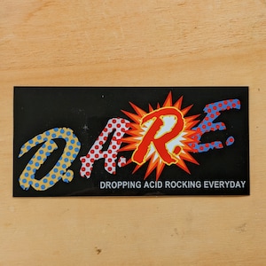 May include: Black rectangular sticker with the word "DARE" in a stylized font. The "D" and "A" are blue with yellow polka dots, the "R" is red with white polka dots, and the "E" is blue with red polka dots. The text "DROPPING ACID ROCKING EVERYDAY" is below.
