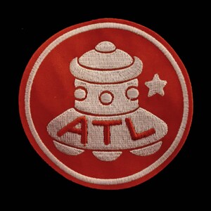 May include: Embroidered patch featuring a red circle with a white border. Inside, a white UFO-like figure with the letters "ATL" in red, and a white star. The patch is on a black background.
