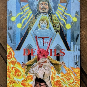 May include: A rectangular poster featuring a colorful illustration for the film The Devils. The artwork displays a central figure in a white habit, a red figure suspended by chains, and a man with a mustache above, all set against a backdrop of blue architectural structures and bright orange flames.
