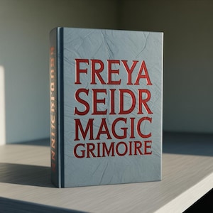 Puede incluir: Un libro de tapa dura titulado "FREYA SEIDR MAGIC GRIMOIRE" en letras rojas sobre una cubierta azul claro texturizada. El libro está sobre una estantería de color claro. El lomo dice "HEIDRUNN JONN".