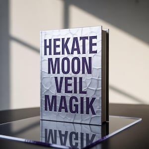 May include: A hardcover book with the title "HEKATE MOON VEIL MAGIK" in purple lettering. The cover has a textured, silver finish. The book is standing upright on a reflective surface, with its reflection visible.