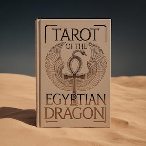 Könnte beinhalten: Ein Hardcover-Buch mit dem Titel "TAROT OF THE EGYPTIAN DRAGON" steht auf einer sandigen Oberfläche. Das Cover zeigt eine goldene Illustration eines Drachen, Flügel und ein Ankh-Symbol. Der Buchrücken ist sichtbar.