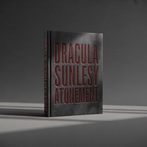 May include: A hardcover book titled "DRACULA SUNLESY ATONEMENT" in red lettering on a dark gray background. The book's spine also features text. The book is standing upright, with shadows cast across it.