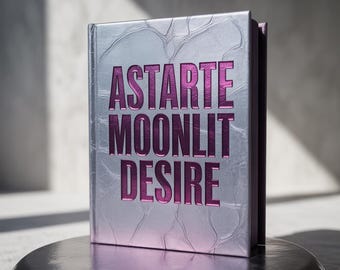 May include: A silver book with the words "ASTARTE MOONLIT DESIRE" in purple lettering. The book is standing on a silver, reflective surface. The cover has a textured, patterned design.