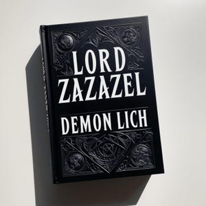 Può includere: Un libro rilegato nero con un designo in rilievo intricato sulla copertina. Il titolo "LORD ZAZAZEL DEMON LICH" è stampato in bianco.