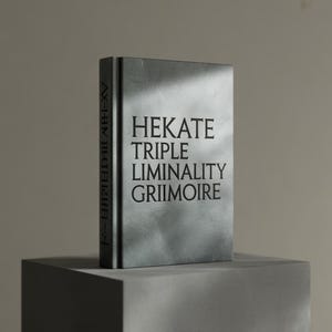 Peut inclure: Un livre gris intitulé "HEKATE TRIPLE LIMINALITY GRIMOIRE" est posé verticalement sur un bloc gris. La tranche du livre affiche du texte vertical. Le livre a un aspect texturé et nuageux.