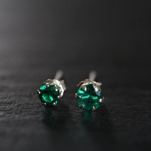 Puede incluir: Un par de pendientes de botón con piedras preciosas de color verde brillante engastadas en plata. Los pendientes son redondos y facetados, reflejando la luz. El fondo es una superficie oscura y texturizada.
