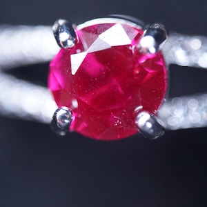 Könnte beinhalten: Nahaufnahme eines silbernen Rings mit einem facettierten Rubin-Edelstein. Der Rubin hat eine tiefe, satte rote Farbe und wird von vier silbernen Krappen gehalten. Der Ring ist geteilt und mit kleinen, funkelnden Steinen besetzt.