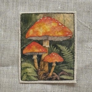 May include: A rectangular patch featuring a watercolor painting of three mushrooms. The mushrooms have orange and yellow caps with white spots, set against a backdrop of green ferns and foliage. The patch is bordered by a cream-colored fabric.