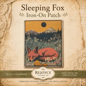 May include: A rectangular iron-on patch featuring an embroidered scene of a red fox sleeping in a field of wildflowers. The background displays a dark forest, snow-capped mountains, and a black sun against a golden sky. Text reads Sleeping Fox.