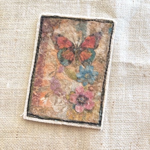 May include: Rectangular fabric patch featuring a butterfly with orange and teal wings, and colorful flowers. The patch has a beige border with black stitching, set against a textured beige background.
