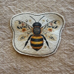 May include: A decorative bee-shaped patch with a black outline. The bee has a black and yellow striped body, black legs, and wings adorned with floral patterns in shades of orange and yellow. The patch is on a beige background.