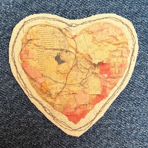 May include: A heart-shaped fabric patch featuring a collage of vintage paper textures in shades of cream, yellow, and red. The patch is outlined with a dark, irregular hand-stitched thread pattern that crisscrosses over the surface of the heart.