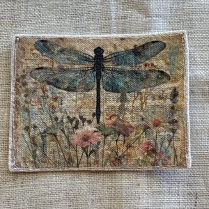 May include: A rectangular fabric patch featuring a dragonfly design. The dragonfly has teal wings and a black body, set against a backdrop of wildflowers in shades of pink, green, and brown. The patch is bordered with a white stitched edge.
