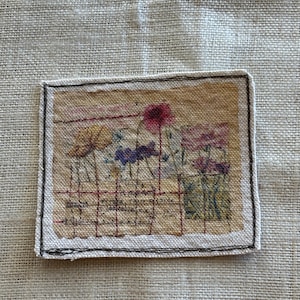 May include: A rectangular textile art piece featuring a floral design with various colors, including yellow, purple, and pink. The artwork has a hand-stitched appearance with a black border and text at the bottom. The background is a neutral linen fabric.