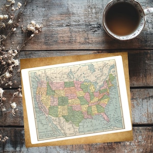 May include: A vintage map of the United States, featuring a detailed depiction of each state in various pastel colors. The map is set within a white border and a brown frame, resting on a rustic wooden surface. A cup of tea sits nearby.