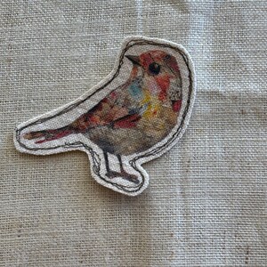 May include: A fabric patch in the shape of a bird. The bird has a colorful design with red, brown, yellow, and blue hues. The patch is outlined with a black stitched border and is set against a neutral linen background.