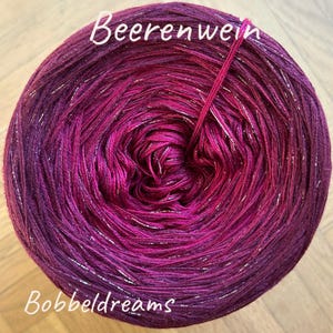 Könnte beinhalten: Ein Garnknäuel in Lila- und Magentatönen mit eingewebten silbernen Metallicfäden. Das Garn ist mit "Beerenwein" und "Bobbeldreams" beschriftet. Das Garn ist in einem Farbverlaufmuster gewickelt, das von Dunkellila zu hellem Magenta übergeht.