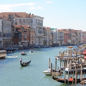 May include: A view of a canal in Venice, Italy, with a gondola in the foreground and several buildings lining the canal. The buildings are a mix of brick and stucco, and some have balconies. The water is calm and reflects the sky.
