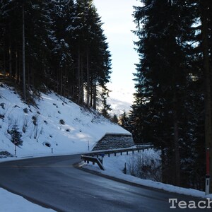 Puede incluir: Un camino sinuoso cubierto de nieve atraviesa un denso bosque de árboles de hoja perenne. El camino está bordeado por bancos cubiertos de nieve y un muro de piedra.