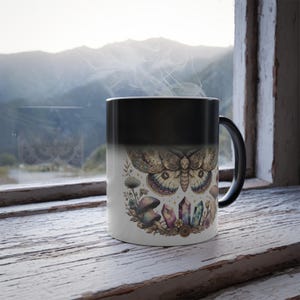 Puede incluir: Taza que cambia de color, negra y blanca, con una ilustración detallada de una polilla, setas, cristales y flores. La taza está colocada junto a una ventana con vistas a la montaña, con vapor saliendo de la taza.